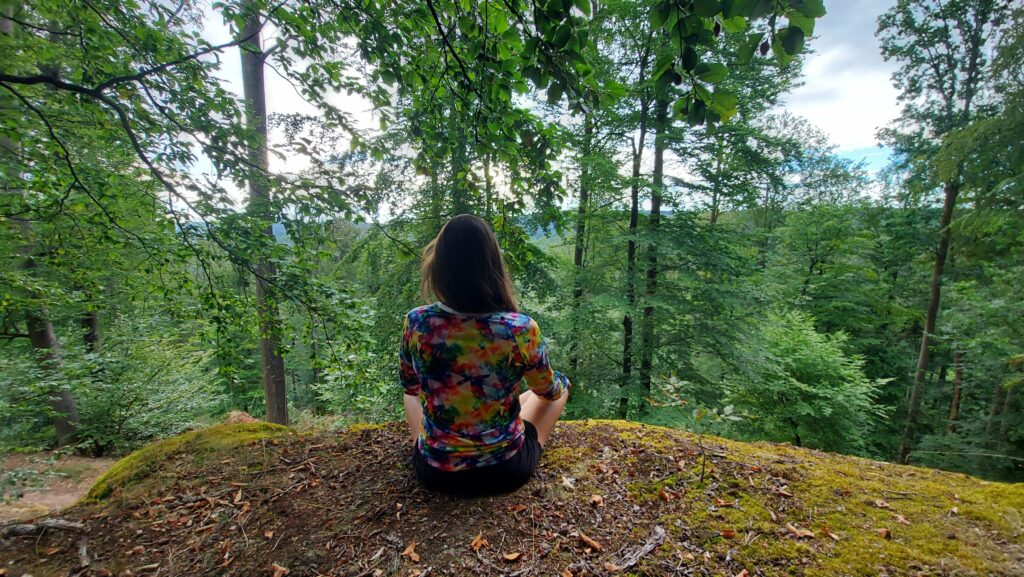 Cindy assise de dos en tailleur, en plein été, face à un massif forestier verdoyant